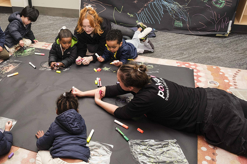 A Cubbyhood production photo. Children and Polyglot artists sit and lie on the ground, drawing together on a large piece of black paper. Another shared drawing hangs on the wall next to them. Photo: Suzanne Phoenix