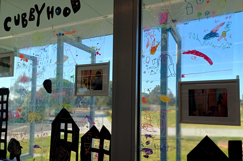 A behind-the-scenes photo of Cubbyhood: A Window Exhibition. Children’s drawings and cardboard house cut-outs are displayed on a window, alongside some framed production photos by Suzanne Phoenix. Photo: Ashlee Hughes