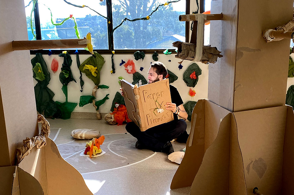 A behind-the-scenes in-school Paper Planet photo. A Polyglot artist sits among tall brown cardboard trees, reading the large Paper Planet book. Photo: Lexie Wood