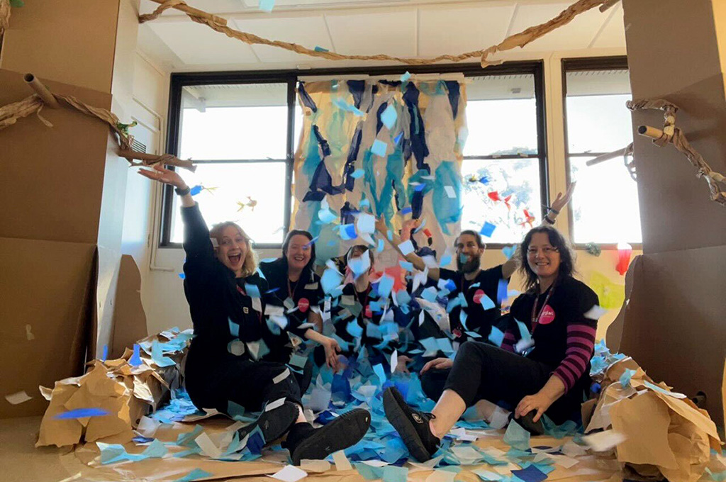 A behind-the-scenes in-school Paper Planet photo. Four Polyglot artists and a Project Manager sit in the waterfall, amidst a shower of blue tissue paper water. Photo: Lexie Wood