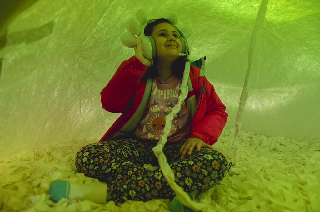 A Voice Lab production photo. A smiling child sits cross-legged in a soft white dome with a white textured floor, illuminated with green light. They wear an intricate set of embellished white headphones which are attached to the space with a long cord. Photo: Theresa Harrison