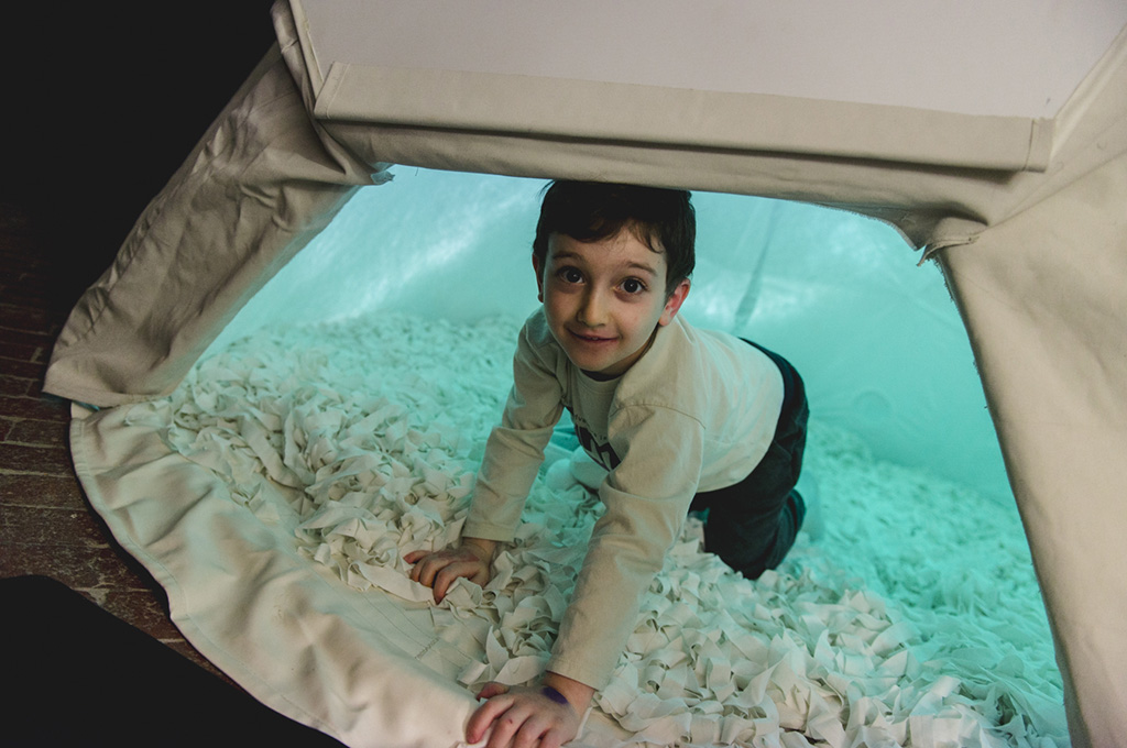 A Voice Lab production photo. A child emerges from a small white dome, smiling at the camera. The interior of the dome is illuminated with blue light. Photo: Theresa Harrison