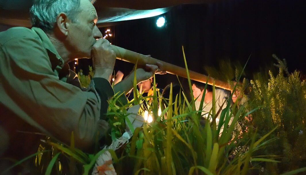 A When the World Turns schools production photo. A Polyglot artist in a khaki shirt uses a long cardboard tube to make sounds, next to a group of young people. They are in a darkened space, surrounded by plants. Photo: Rainbow Sweeny