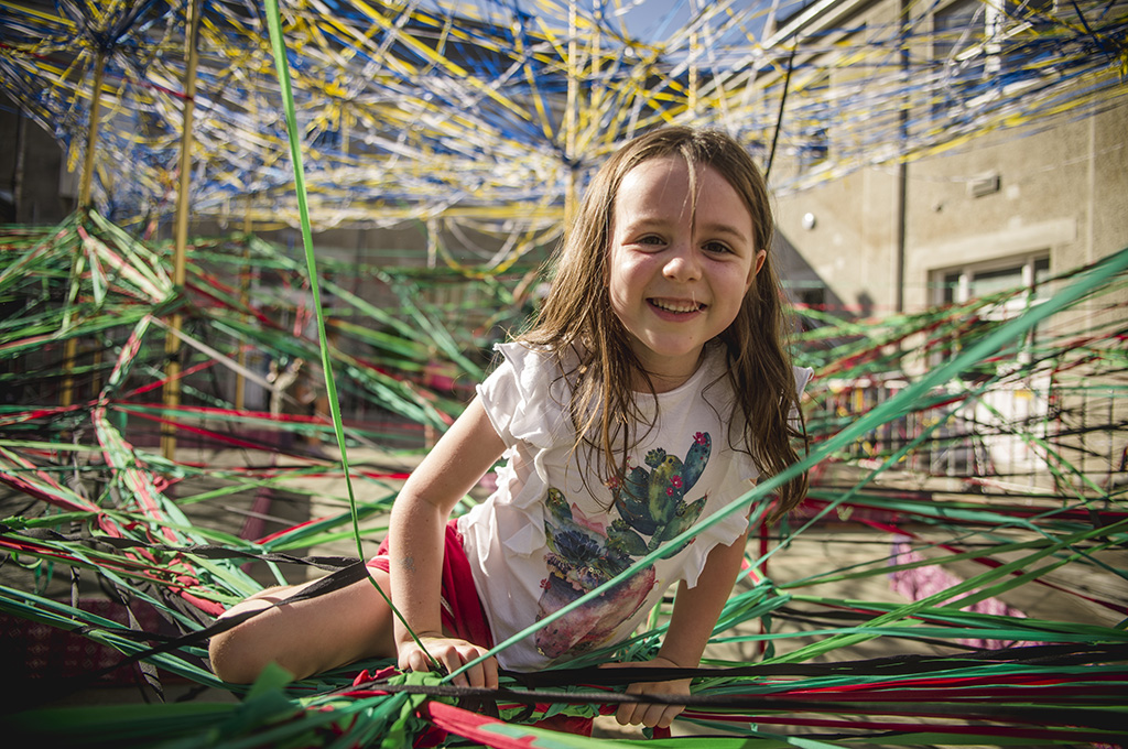 A Tangle production photo. A child climbs among hundreds of strands of coloured elastic woven through tall golden poles in a heritage courtyard. They smile at the camera. Photo: Theresa Harrison.