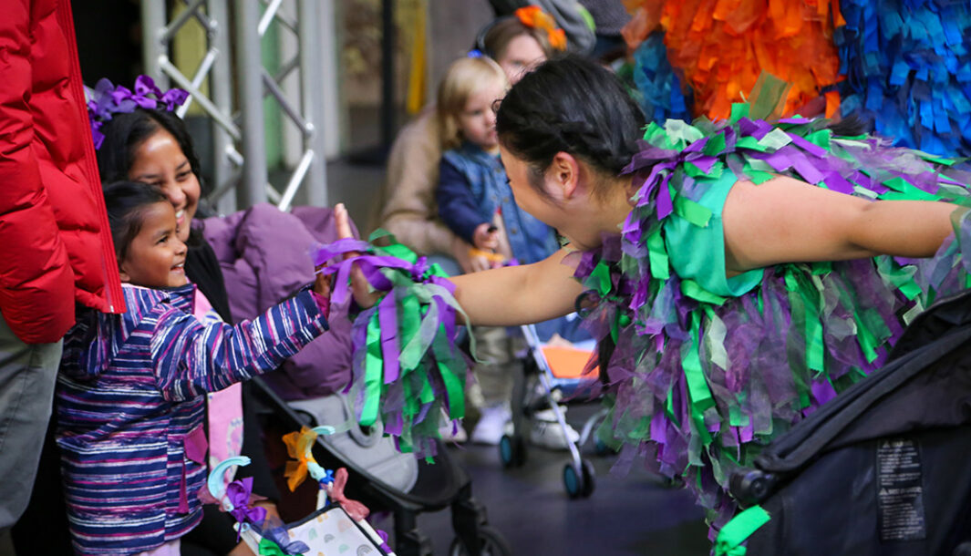 A Pram People production photo. A Polyglot artist in a purple and green costume and a small child reach towards each other. The child's adult wears headphones decorated with purple ribbon, and smiles at the exchange. Photo: Kenny Waite, Abbotsford Convent Foundation.