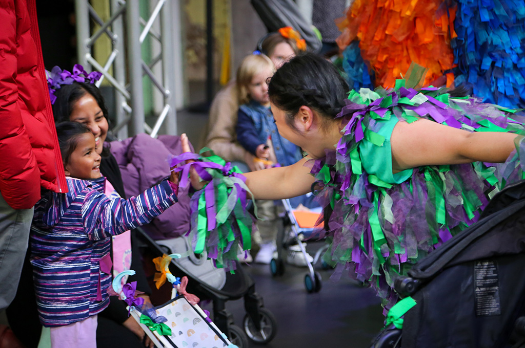 A Pram People production photo. A Polyglot artist in a purple and green costume and a small child reach towards each other. The child's adult wears headphones decorated with purple ribbon, and smiles at the exchange. Photo: Kenny Waite, Abbotsford Convent Foundation.