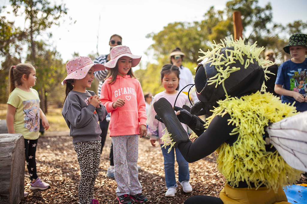 A Bees production photo. A Polyglot artist in an intricate black and yellow Bee costume engages with three children. Adults and other children look on. Photo: Royal Botanic Gardens Victoria.