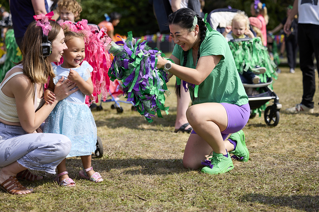 A Pram People production photo. A Polyglot artist in a purple and green costume engages with a small child and their adult, who is wearing headphones. Small children in prams decorated with brightly coloured ribbon are visible in the background. Photo: David Levene, GDIF