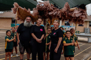 Totems production photo, Wailwan Country/Warren: Belinda Bell, 2024. Students, artists Blayne, Tamara, and Polyglot Producer Julie, stand in the playground, smiling. The students hold up the large Maliyan (Wedgetail eagle) puppet, made from paper and bamboo.