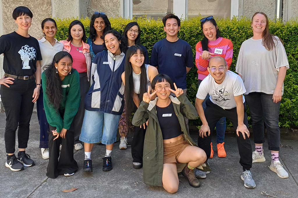 CAAP Artistic Director Tessa Leong, Polyglot Artistic Director Cat Sewell and 11 artists smile for the camera. They are outside, in a heritage courtyard. 