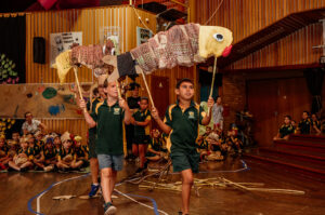Totems production photo, Wailwan Country/Warren: Belinda Bell, 2024. Students in green and gold uniforms manoeuvre their Guduu (Murray cod) puppets, made from paper and bamboo, into the performance space in the school hall. Other students sit on the side, watching.