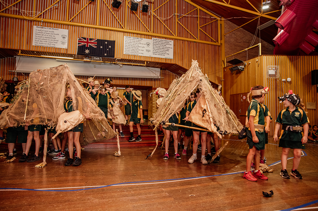 A Totems production photo. Groups of students operate two large Dhigarrbila (echidna) puppets, made from paper, hessian and bamboo. Others in paper costumes gather around them. Photo: Belinda Bell, 2024