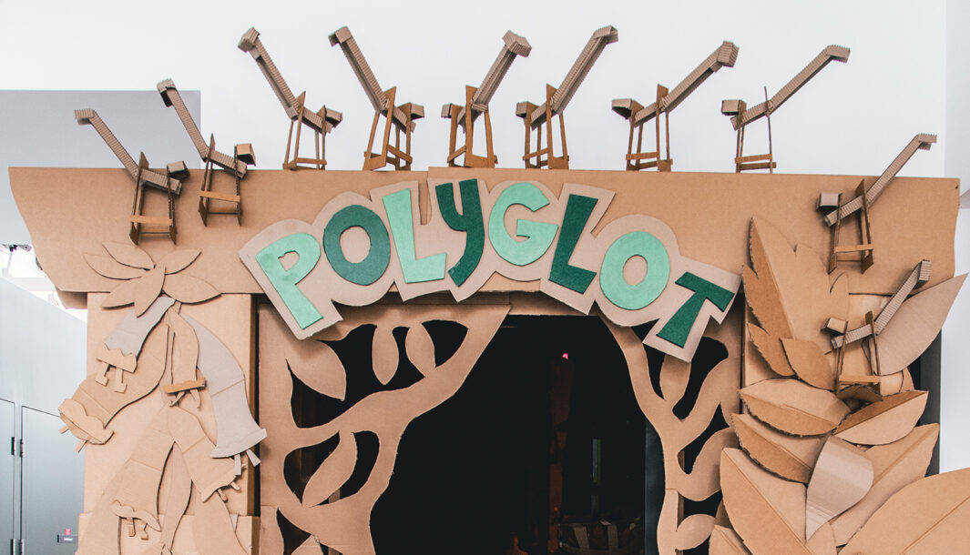 Paper Planet production photo: Jacinta Keefe, Footscray Community Arts. An intricate, handmade cardboard arch, featuring cardboard container cranes, leaves and gumnuts, opens into a dark performance space. Green letters spelling 'Polyglot' curve across the top.