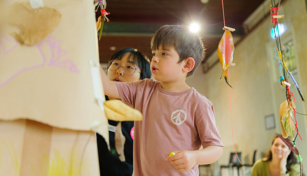 Art Tree production photo. A child and a Polyglot artist use markers to add to a brown paper tree. Colourful, decorated leaves and twigs are suspended with yarn. The child’s adult looks on. Photo: Sarah Walker