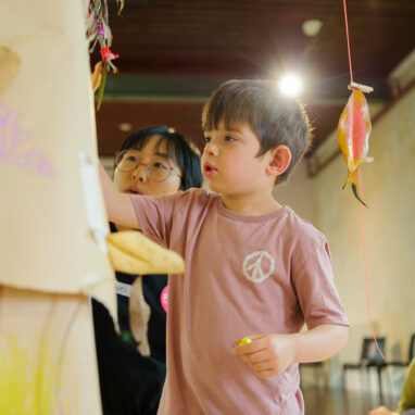 Art Tree production photo. A child and a Polyglot artist use markers to add to a brown paper tree. Colourful, decorated leaves and twigs are suspended with yarn. The child’s adult looks on. Photo: Sarah Walker