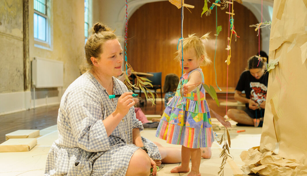 An Art Tree production photo. A small child reaches towards a branch suspended from a large tree with colourful yarn. They are watched by their adult, who holds a marker. Photo: Sarah Walker