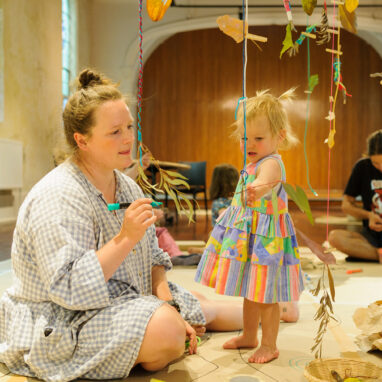 An Art Tree production photo. A small child reaches towards a branch suspended from a large tree with colourful yarn. They are watched by their adult, who holds a marker. Photo: Sarah Walker