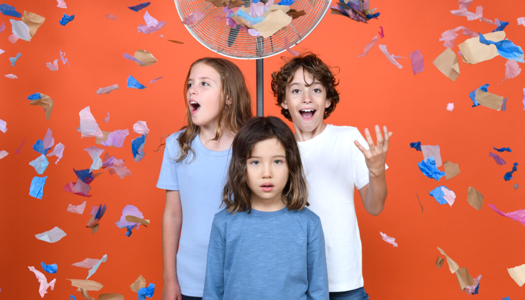 Three children stand against an orange wall, in-front of a fan. There is confetti in the air. Photographer: Sarah Walker.