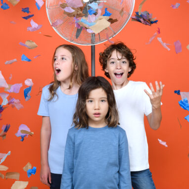 Three children stand against an orange wall, in-front of a fan. There is confetti in the air. Photographer: Sarah Walker.