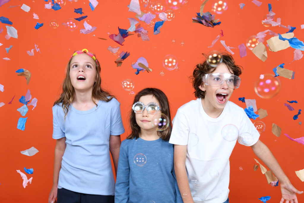 Three children stand against an orange wall, in-front of a fan. There is confetti in the air. Photographer: Sarah Walker.