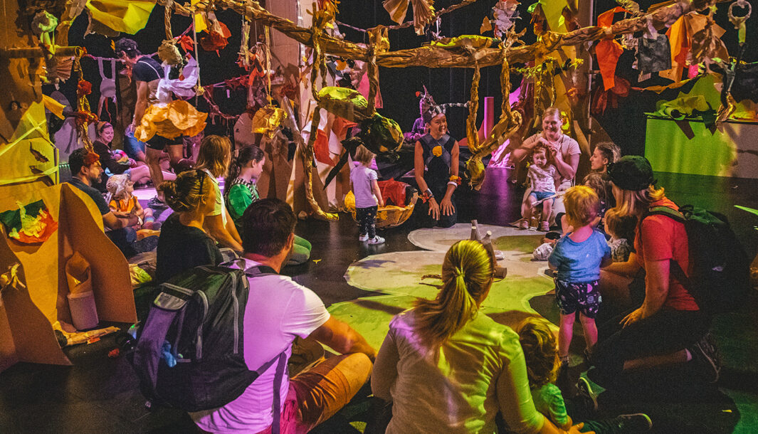 Paper Planet production photo: Jacinta Keefe, Footscray Community Arts. Families and Polyglot artists form a large circle on the floor. They are in a forest of tall cardboard trees, in a darkened space with colourful, theatrical lighting.