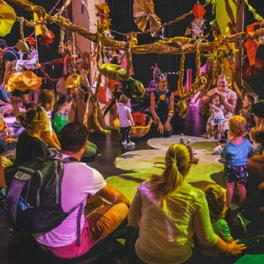 Paper Planet production photo: Jacinta Keefe, Footscray Community Arts. Families and Polyglot artists form a large circle on the floor. They are in a forest of tall cardboard trees, in a darkened space with colourful, theatrical lighting.