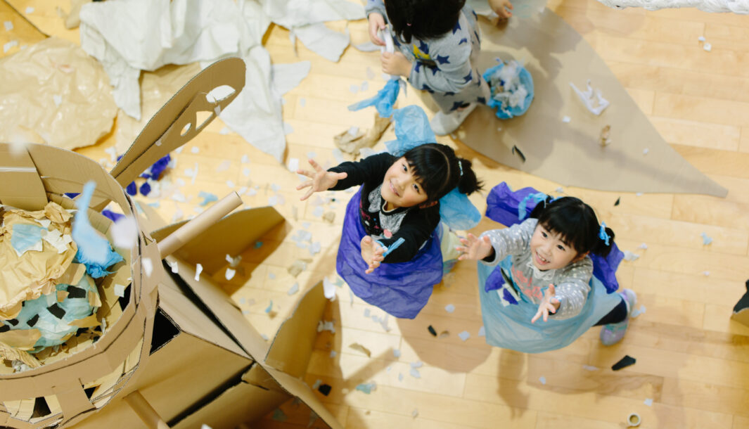 A Paper Planet production photo. Two children in blue look up, their hands wide open as paper falls down. Photo: Ai Ueda