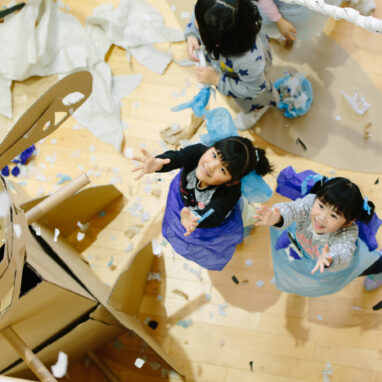 A Paper Planet production photo. Two children in blue look up, their hands wide open as paper falls down. Photo: Ai Ueda