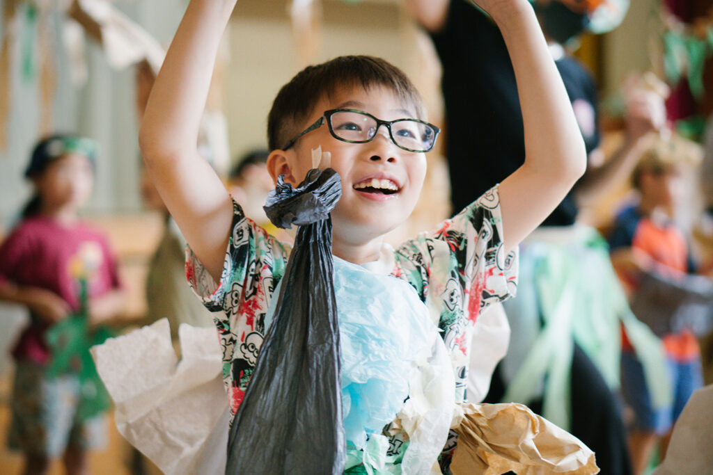 Paper Planet production photo: Ai Ueda. A child wearing glasses, a patterned t-shirt, and a handmade paper costume lifts their arms, moving through the performance space. Polyglot artists and other children are visible behind them.