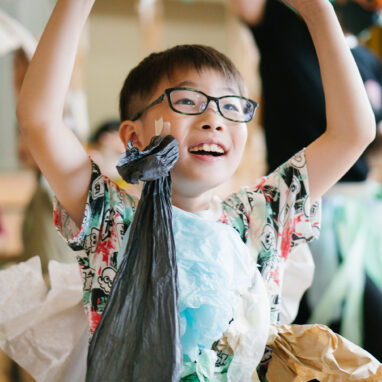 A Paper Planet production photo. A child wearing glasses, a patterned t-shirt, and a handmade paper costume lifts their arms, moving through the performance space. Polyglot artists and other children are visible behind them. Photo: Ai Ueda
