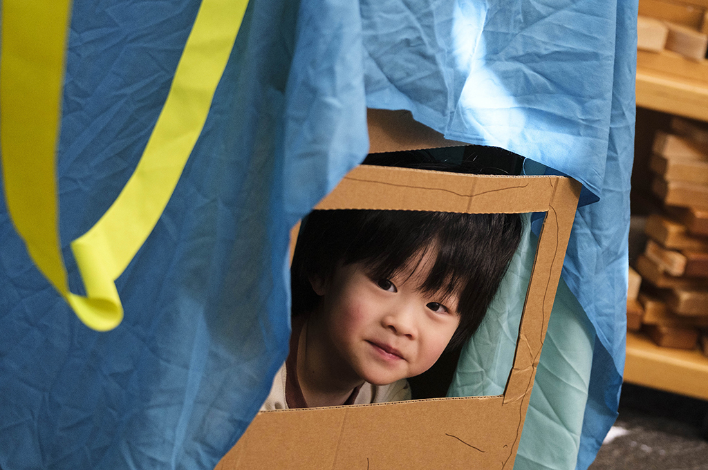Cubbyhood production photo: Suzanne Phoenix. A smiling child with black hair peers through a cardboard window under a blue piece of cloth.