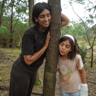 Forest production photo: Claudia Sangiorgi Dalimore. A Polyglot artist and a child lean on either side of a tree, smiling. Another child looks on.