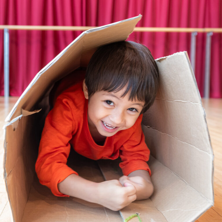 Pass It On production photo: Alvin Ho, Esplanade - Theatres on the Bay, Singapore. A child in an orange jumper climbs through a large cardboard tube. They smile at the camera.