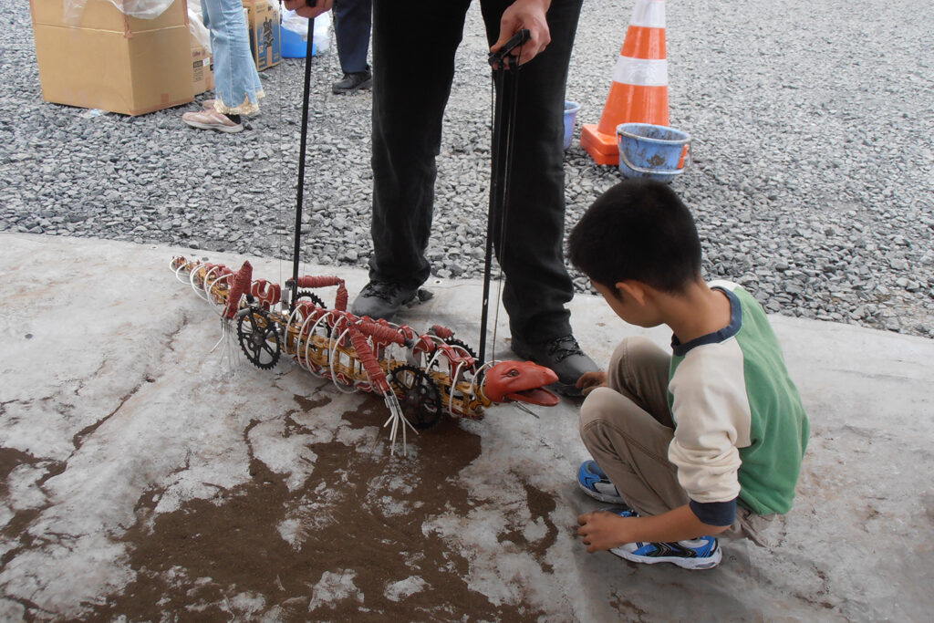 A photo from Polyglot's 2011 engagement in Minami Sanriku. A small child engages kneels on the ground, engaging with a lizard puppet manipulated by a Polyglot artist.