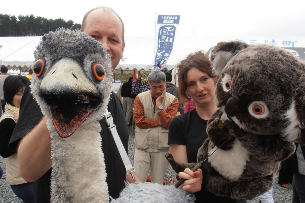 A photo from Polyglot's 2011 engagement in Minami Sanriku. Two Polyglot artists hold large Australian animal puppets (an emu and a koala), smiling at the camera. They are outdoors, in a crowd of people.