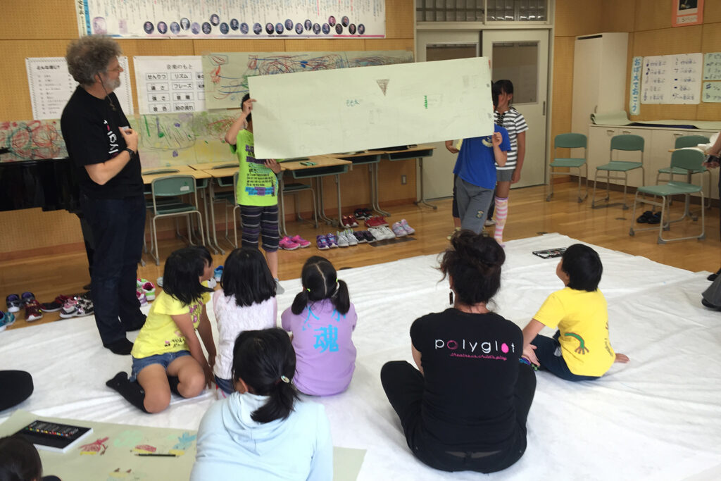 Tusgi Wa? (What Next?) photo: Stefanie Robinson. Children hold up a long drawing in a classroom, with other students and two Polyglot artists looking on.
