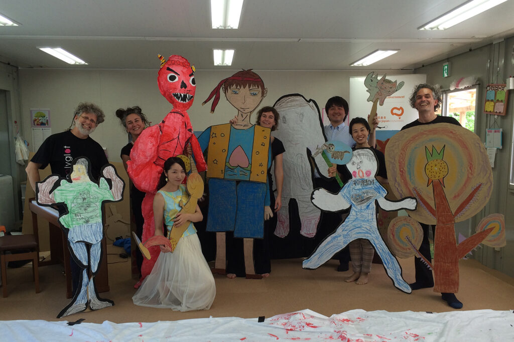 Tusgi Wa? (What Next?) photo. Polyglot and NPO Acchi Cocchi artists hold up large cardboard and paper creations, smiling at the camera.
