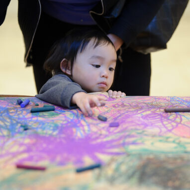 Sound of Drawing production photo: Sarah Walker. A small child stands at a table covered in brown paper, which is filled with shared, abstract drawing. They reach towards coloured pastels.