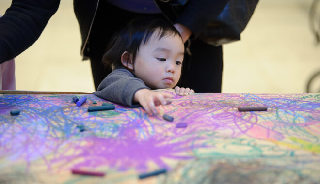 Sound of Drawing production photo: Sarah Walker. A small child stands at a table covered in brown paper, which is filled with shared, abstract drawing. They reach towards coloured pastels.