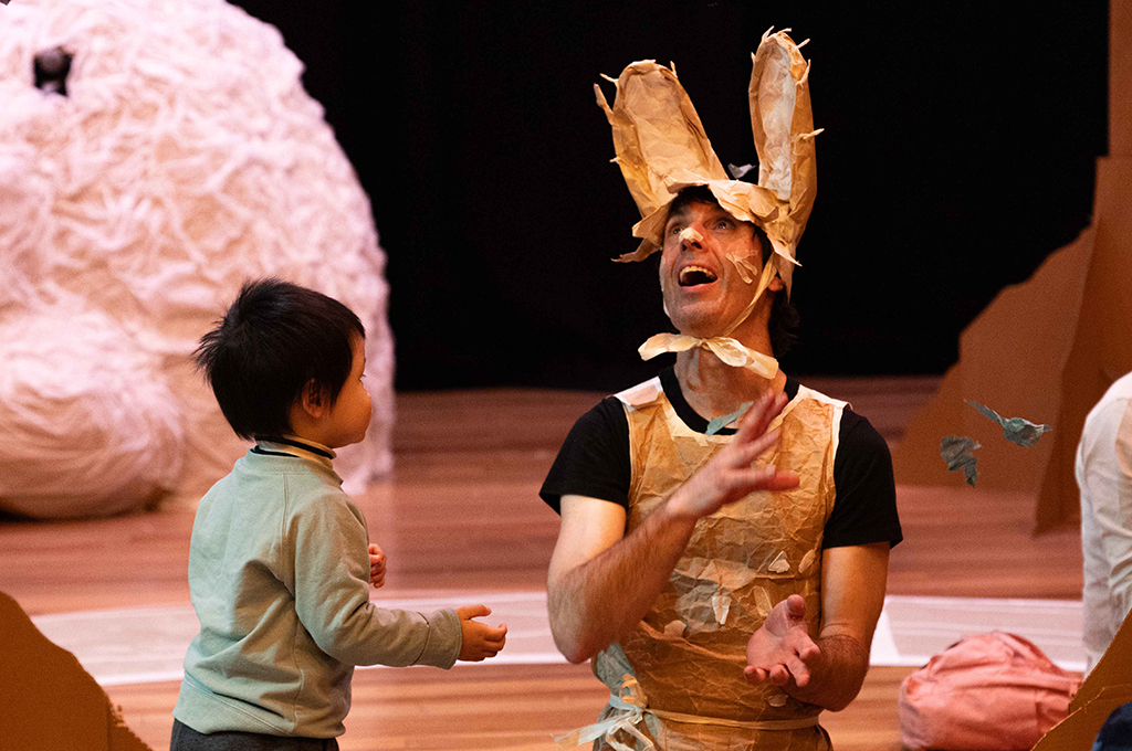 Paper Planet production photo: Katje Ford, Sydney Opera House. A Polyglot artist in an intricate, handmade paper rabbit costume engages with a small child, throwing small pieces of paper into the air. They are indoors, in a theatrically lit space.