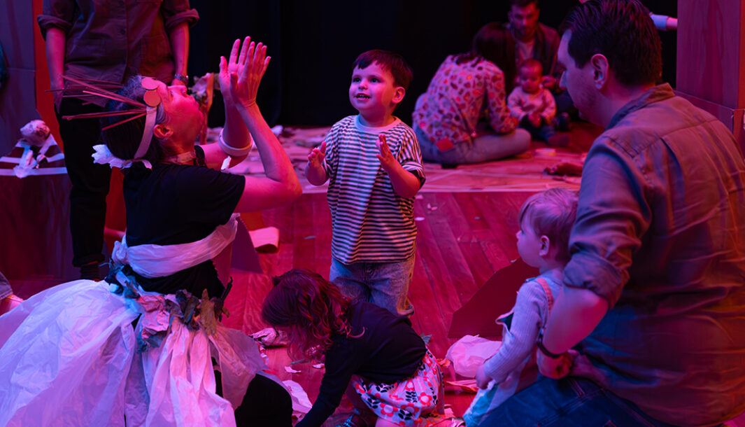 Paper Planet production photo: Katje Ford, Sydney Opera House. A Polyglot artist in an intricate, handmade paper costume engages with a small child, throwing pieces of paper into the air. Other children and adults look on. They are in a darkened, theatrically lit space.