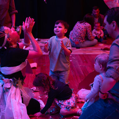 Paper Planet production photo: Katje Ford, Sydney Opera House. A Polyglot artist in an intricate, handmade paper costume engages with a small child, throwing pieces of paper into the air. Other children and adults look on. They are in a darkened, theatrically lit space.