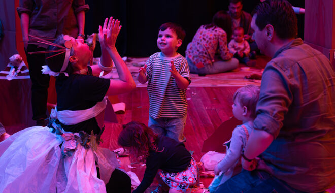 Paper Planet production photo: Katje Ford, Sydney Opera House. A Polyglot artist in an intricate, handmade paper costume engages with a small child, throwing pieces of paper into the air. Other children and adults look on. They are in a darkened, theatrically lit space.