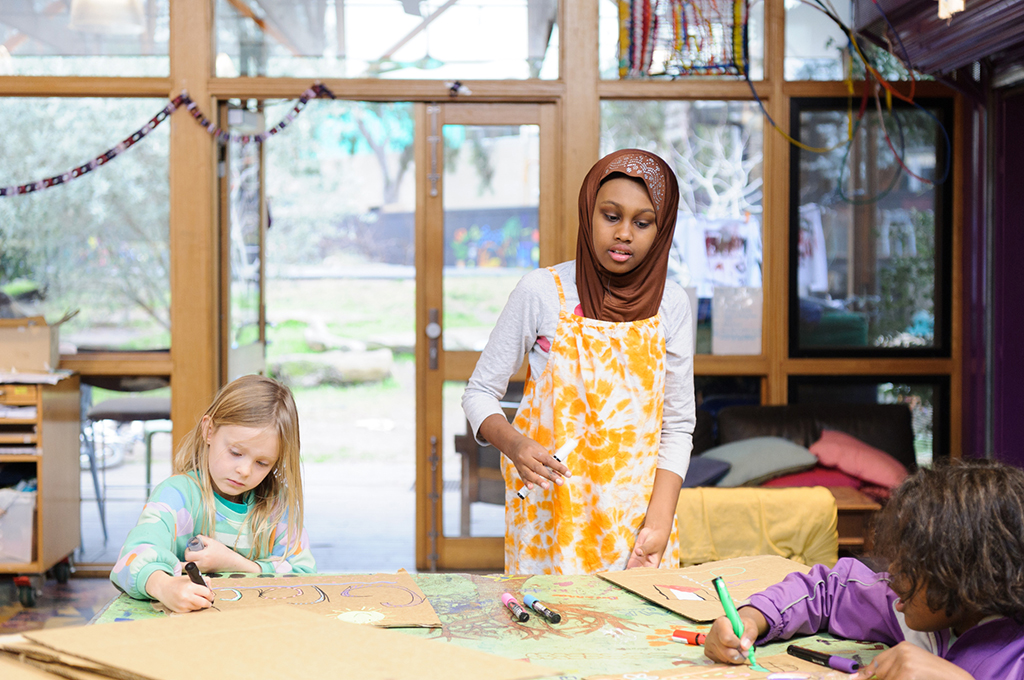 Us. Here. Now. production photo: Sarah Walker. Three children stand and sit around a table, creating and playing with brown paper and markers.