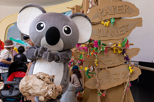 A Paper Planet workshop. A person in a koala costume stands next to a Paper Planet cardboard sign in both Japanese and English. Photo courtesy of DFAT.