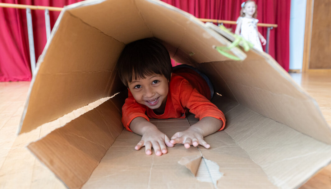 Pass It On production photo: Alvin Ho, Esplanade - Theatres on the Bay, Singapore. A child in an orange jumper climbs through a large cardboard tube. They smile at the camera.