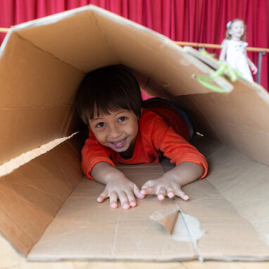 Pass It On production photo: Alvin Ho, Esplanade - Theatres on the Bay, Singapore. A child in an orange jumper climbs through a large cardboard tube. They smile at the camera.