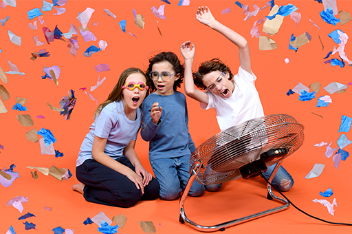 Three children wearing goggles stand against an orange wall. There is confetti and bubbles in the air. Photographer: Sarah Walker.
