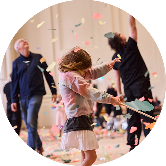 Whirlwind production photo: A child braces for the impact of swirling colourful confetti. An adult and Polyglot artist stand in the background, looking up at the confetti. Photo: Laura May Grogan.