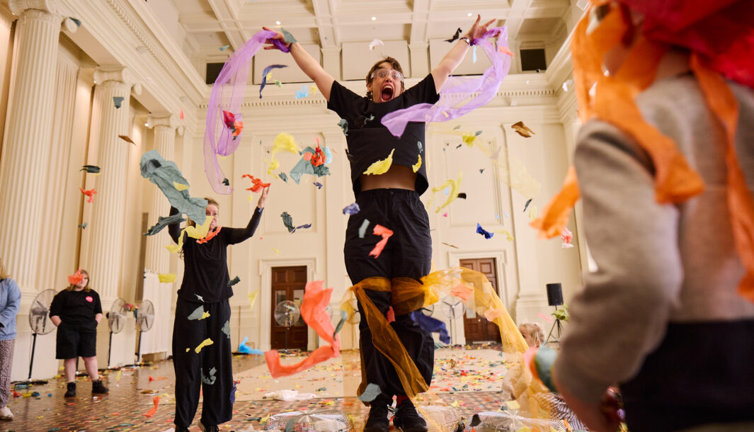 Whirlwind production photo: Laura May Grogan. Two Polyglot artists leap in the air as colourful confetti falls. A child stands in the corner and watches on.